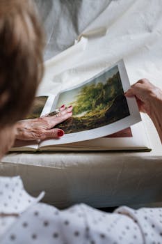 An elderly woman turns pages of a landscape art book at home, exploring detailed illustrations.